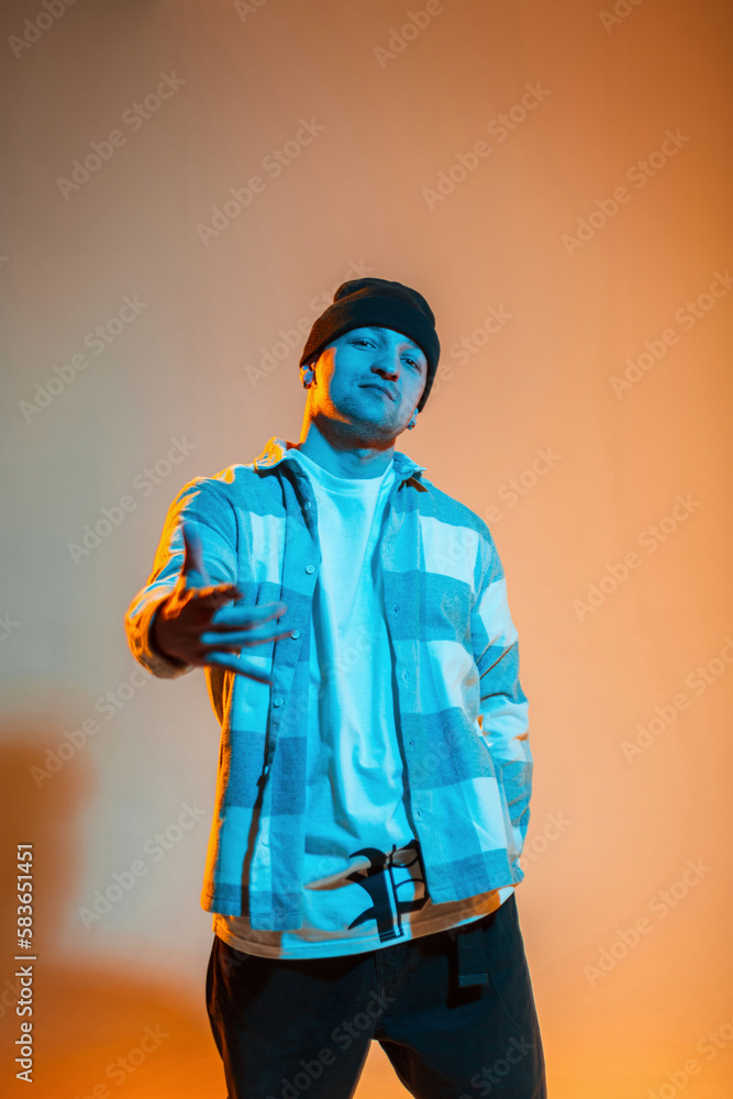 Stylish b-boy dancer guy with hat in a trendy fashion plaid shirt stands on an orange background with neon light Stylish b-boy dancer guy with hat in a trendy fashion plaid shirt stands on an orange background with neon light