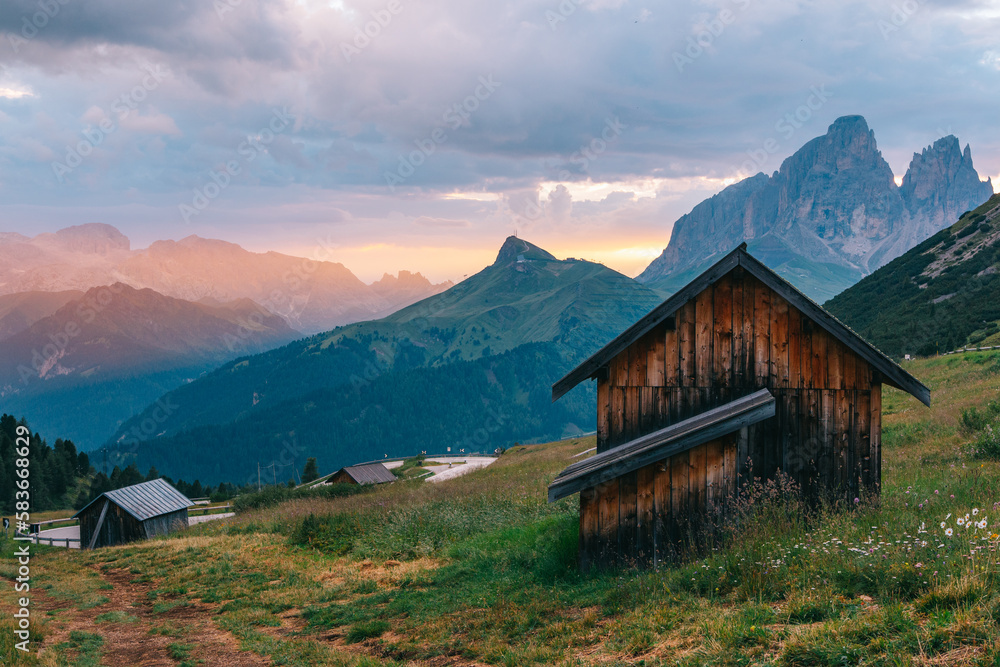 Italian Alps picturesque mountain pass, quiet place of wooden houses surrounded by green meadows. Dawn with warm golden, creating a dreamlike landscape to explore and discover beauty of alpine nature.