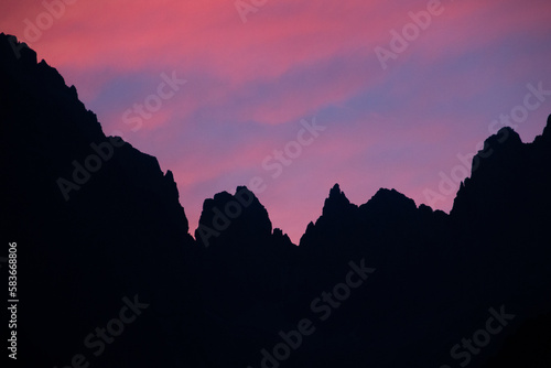 Rugged peaks of the mountain range are silhouetted against a stunning sunset sky, awash with a kaleidoscope of warm and pink colors that create a truly breathtaking vista. Dolomites, Alps, Italy.