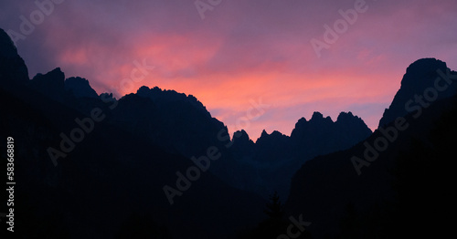 Majestic silhouette of the mountain range stands tall against a sky painted with hues of purple and pink, as the last rays of sunlight peek over the horizon. Italian Dolomites Alps, panoramic view.