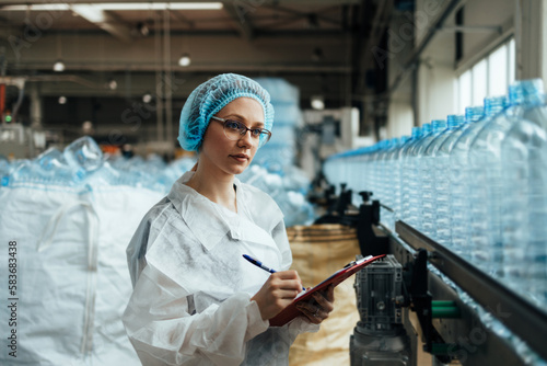 Female worker in protective workwear working in medical supplies research and production factory and checking canisters of distilled water before shipment. Inspection quality control.