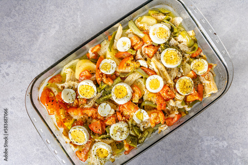 Gomes de sa codfish roasted in olive oil with tomatoes, peppers, onions, boiled eggs and oregano. In a rectangular glass baking dish.