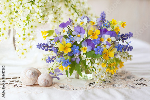 A bouquet of spring blue, yellow, white, violet flowers in a vase on the table. Pansies, forget me nots, primroses, bird cherry, violets, muscari. Postcard, blur, selective focus.