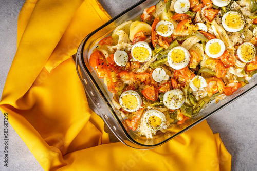 Gomes de sa codfish roasted in olive oil with tomatoes, peppers, onions, boiled eggs and oregano. In a rectangular glass baking dish surrounded by an orange towel.