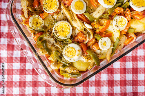 Gomes de sa codfish roasted in olive oil with tomatoes, peppers, onions, boiled eggs and oregano. In a rectangular glass baking dish on a checkered towel.