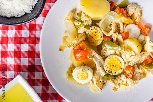 Gomes de sa codfish roasted in olive oil with tomatoes, peppers, onions, boiled eggs and oregano. In a white porcelain dish on a checkered tablecloth with cutlery, rice and olive oil.