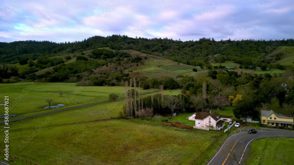 Car drives by country houses by fields in green California landscape at dawn