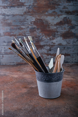 In a tin bucket there are many different in shape and size brushes for painting with natural bristles in oils, palette knives are blurry, in the sunlight in the corner of the artist's studio