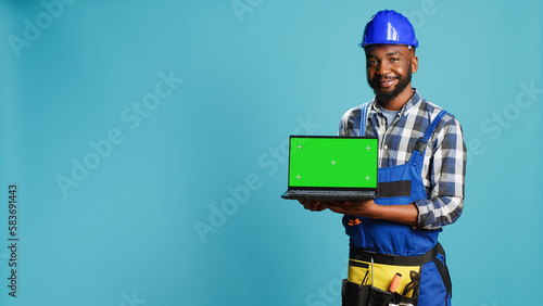 Smiling craftsman showing laptop with green screen on camera, advertising modern isolated display on pc. Young builder contractor holding computer with blank chroma key copyspace screen. © DC Studio