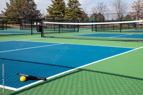 View of a pickleball complex with a paddle and yellow ball on blue and green courts beside a playground in a suburban park in early spring. © pics721