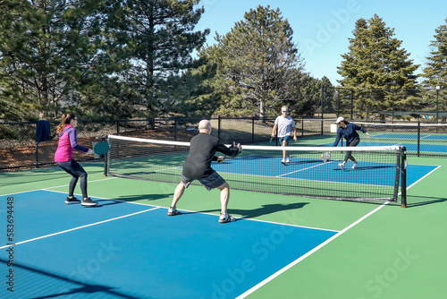 A competitive doubles game of pickleball with a group of men and women on a blue and green court in spring.