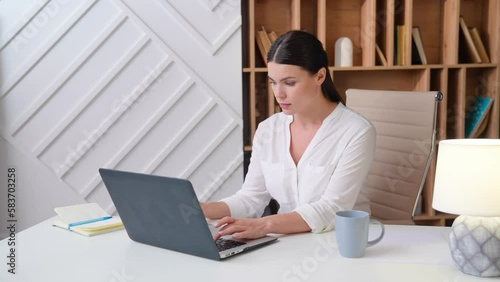 Young business woman boss works on modern laptop focused doing research in office. Young female freelancer worker in white blouse concentrated sitting at table at home typing on laptop