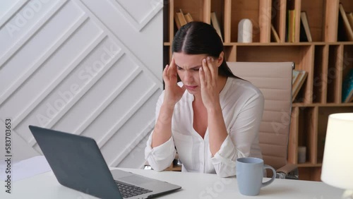 Young woman worker sitting at the table typing on the laptop taking a break tired rubbing her temples headache in the office at home camera is approaching