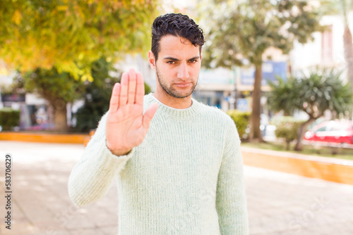 young hispanic man looking serious, stern, displeased and angry showing open palm making stop gesture