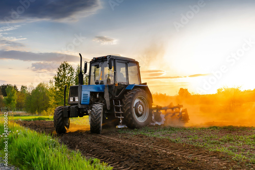 Fototapeta Modern blue tractor machinery plowing agricultural field meadow at farm at spring autumn during sunset
