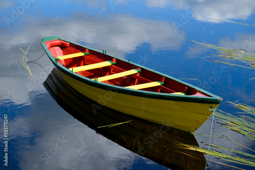 A small yellow wooden row boat with green trim and a red interior floats on calm water with reeds in the water. The boat is tied on with green rope. The boat's reflections are seen in the water.