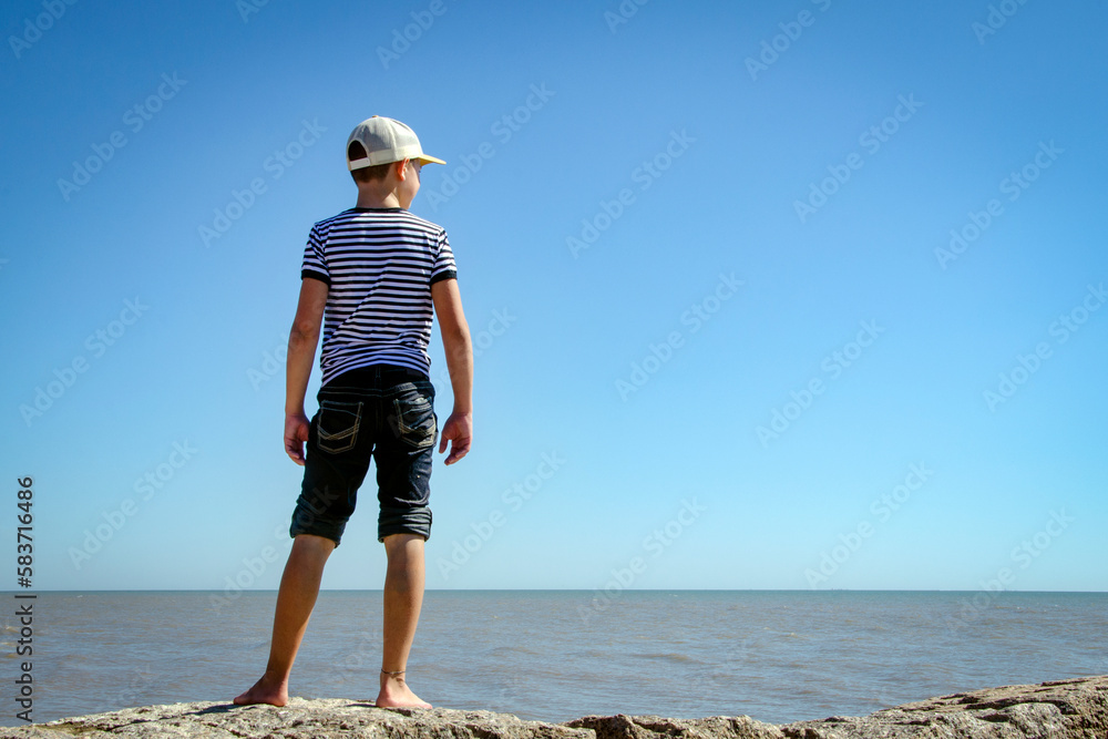 Young preteen boy standing on sunny beach wearing jeans and hat