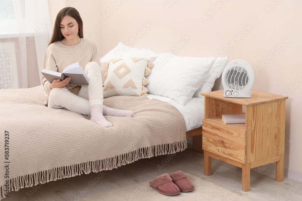 Woman reading book near portable electric fan heater in bedroom