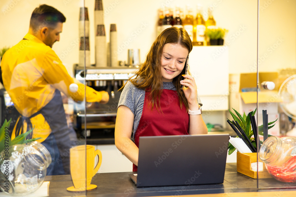 Female cashier with good mood making finance report at end in working day while talking with friends on modern smartphone about meeting after job standing near coffee machine at bar with modern laptop