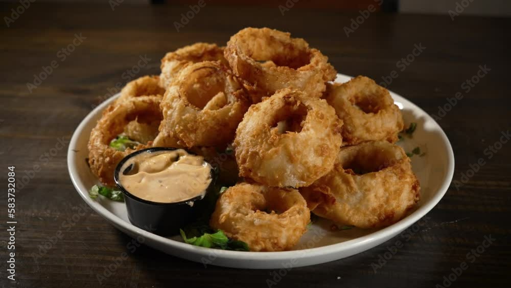 Large deep fried onion rings piled high on white plate with sauce, slider 4K