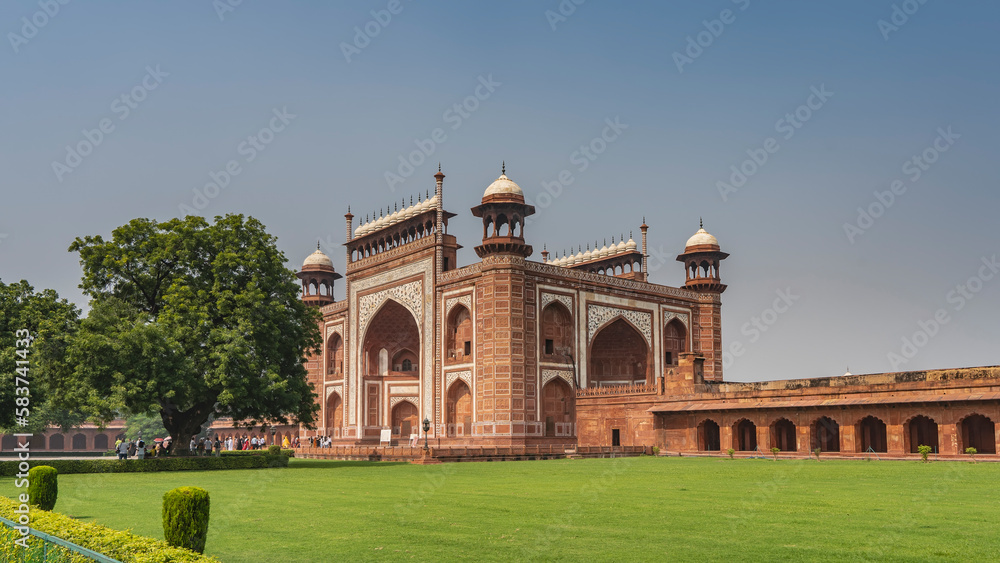 The building in the Taj Mahal complex is built of red sandstone and white marble. Visible arches, domes, spires, gallery. A lawn with green grass. Blue sky. India. Agra