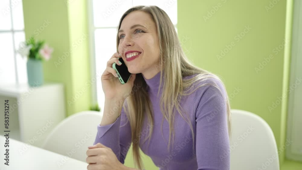 Young blonde woman talking on smartphone sitting on table at home Young blonde woman talking on smartphone sitting on table at home