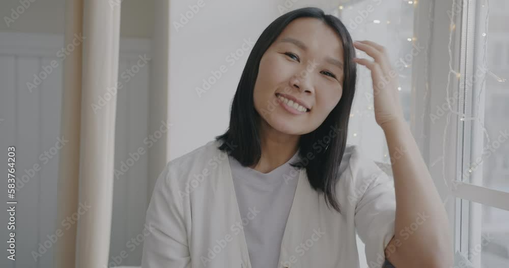 Slow motion portrait of beautiful Asian lady smiling and looking at camera at home with window in background. People and positive emotion concept. Slow motion portrait of beautiful Asian lady smiling and looking at camera at home with window in background. People and positive emotion concept.