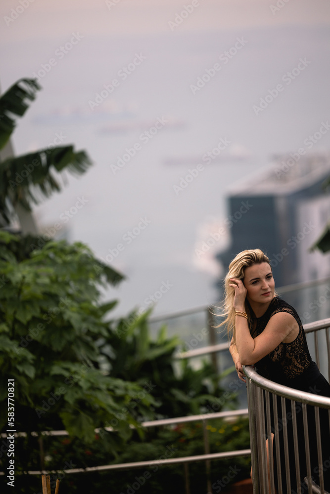 A beautiful Caucasian blonde girl in a luxurious black dress is standing against the backdrop of a park in a modern city. Greenery and concrete in the city of the future. Black dress concept