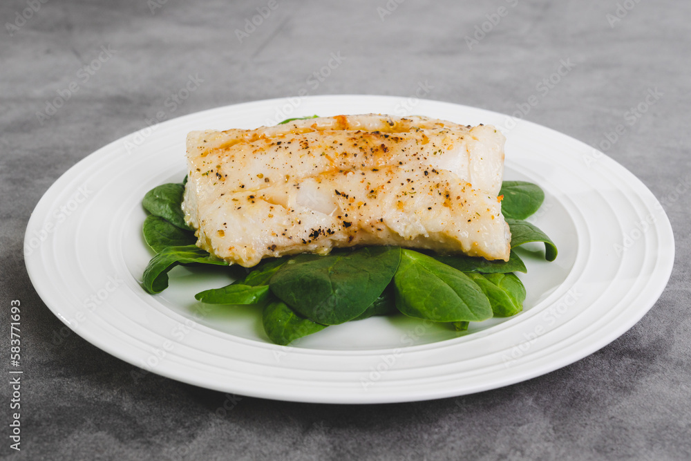 Cod fillet baked with garlic butter sauce served with fresh spinach on a white plate close-up on the kitchen table Cod fillet baked with garlic butter sauce served with fresh spinach on a white plate close-up on the kitchen table