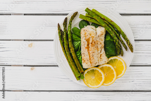 Cod fillet baked with garlic butter sauce served with fresh spinach and roasted asparagus on a white plate close-up
