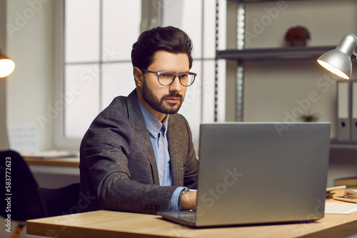 Businessman working in the office. Serious, focused young man in a stylish suit and eyeglasses sitting at his desk, using his laptop computer, doing research, and preparing a business presentation