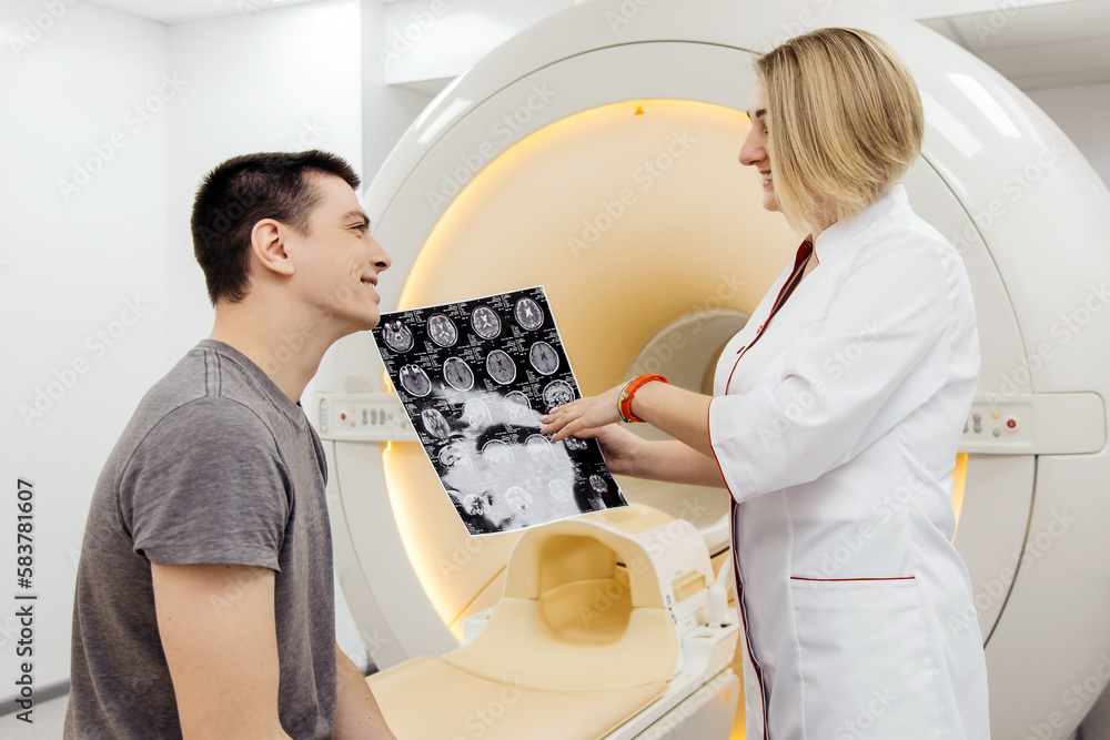 Female doctor radiologist explains good results of CT scan to patient, shows picture with images, observes and analyzes CT scan in modern clinic next to modern CT scanner Female doctor radiologist explains good results of CT scan to patient, shows picture with images, observes and analyzes CT scan in modern clinic next to modern CT scanner