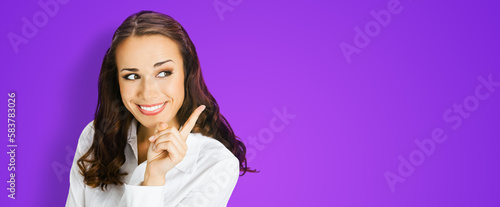 Young happy smiling woman in confident blouse, showing pointing finger aside at copy space. Business ad concept. Brunette businesswoman, isolated violet purple background. Wide image. © vgstudio