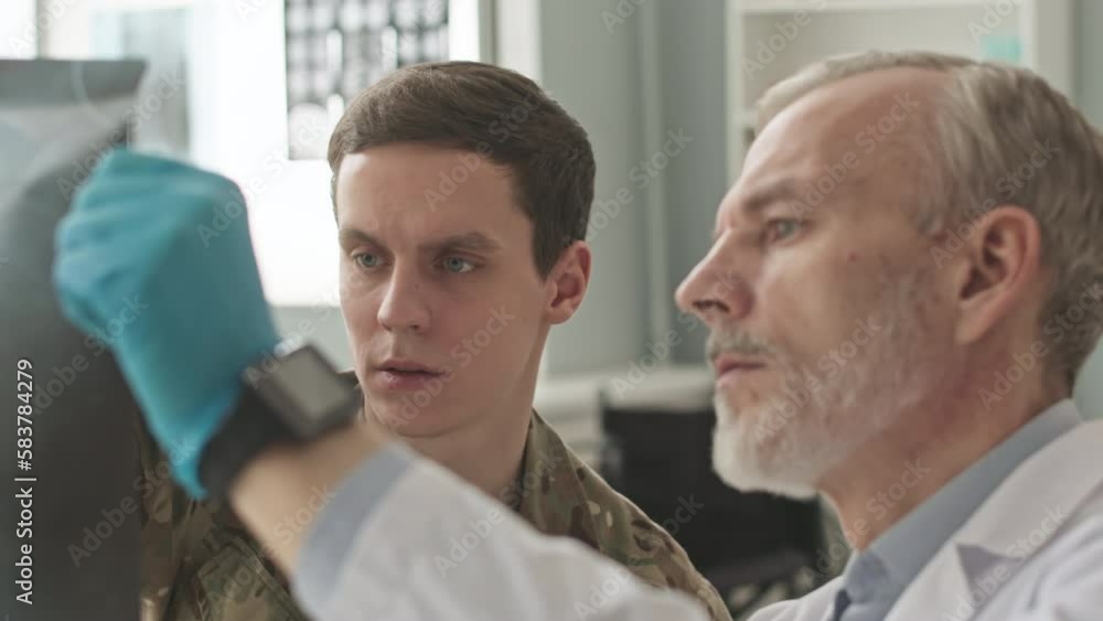 Medium closeup of military doctor showing x-ray image to young army officer in camouflage uniform having checkup appointment at hospital Medium closeup of military doctor showing x-ray image to young army officer in camouflage uniform having checkup appointment at hospital