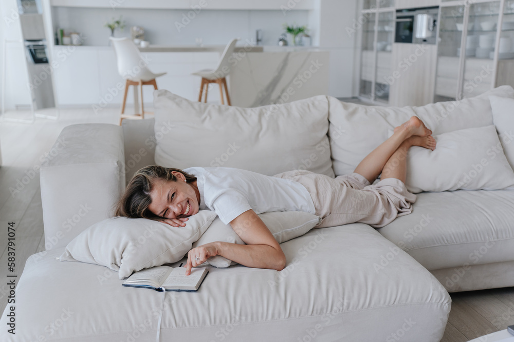Laughing caucasian girl in casual laying on cozy couch points at diary, completed all tasks, relaxing home on vacation. Cheerful American student girl enjoying vacation. Cozy home. Laughing caucasian girl in casual laying on cozy couch points at diary, completed all tasks, relaxing home on vacation. Cheerful American student girl enjoying vacation. Cozy home.