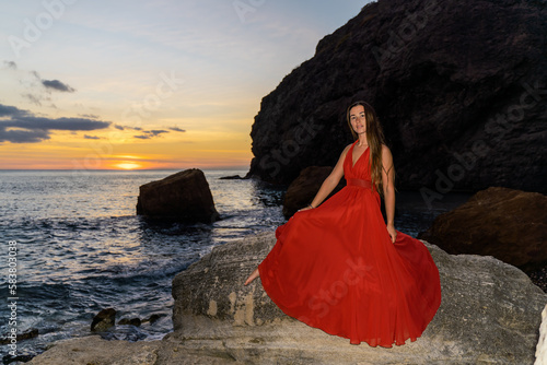 Woman in a red flying dress on the ocean or on the sea beach against the backdrop of the sunset sky.