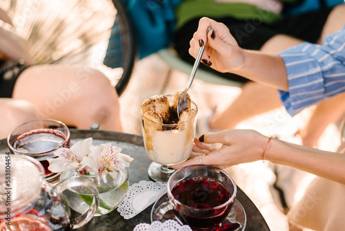 Summer cafe terrace. A girl with a spoon in her hands takes tiramisu dessert from a glass..On the table are glass cups with tea, a teapot and white flowers in a vase.