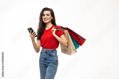 Online shopping. Beautiful young woman holding shopping bags and using her phone with smile while standing against white background