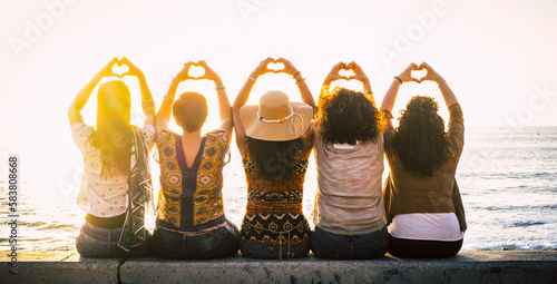 Fotografie Group of women outdoor enjoying leisure and travel summer holiday together in friendship and love sitting against a sunset on the ocean and doing heart sig with raised up arms and hands