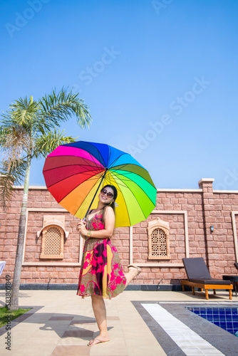 Beautiful young indian woman wearing sunglasses holding rainbow color umbrella in hot summer day.