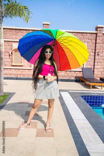 Beautiful happy young indian girl wearing sunglasses holding rainbow color umbrella in hot summer day. Vacation and holidays.