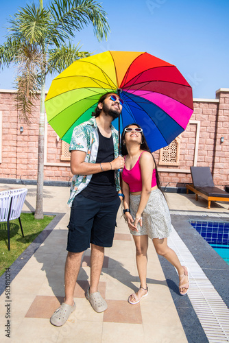 Young indian couple wearing sunglasses holding rainbow color umbrella in hot sunny day. Summer Vacation and holidays.