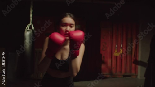 Active motivated sporty fit pretty Asian woman in boxing gloves practicing shadowboxing training, improving punching techniques and skills, throwing jab and hook punch in garage gym