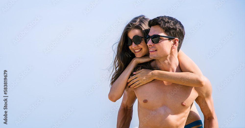 Portrait of beautiful caucasian woman with man wearing sunglasses swim suit piggy back on beach. Young couple enjoy honeymoon after marriage sea. Happy casual lover hold at the tropical beach Portrait of beautiful caucasian woman with man wearing sunglasses swim suit piggy back on beach. Young couple enjoy honeymoon after marriage sea. Happy casual lover hold at the tropical beach