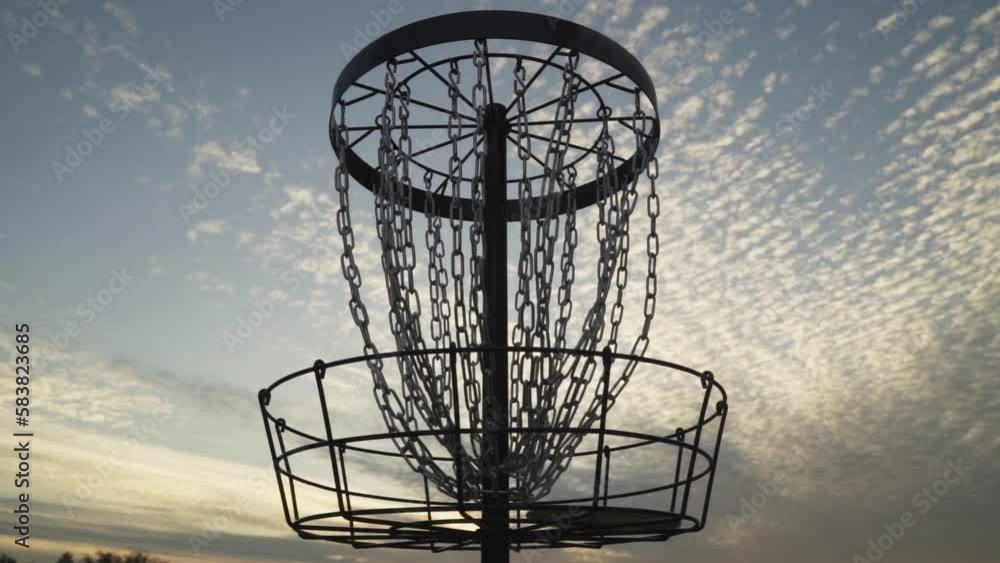 Disc Golf Basket Silhouetted by Evening Sky While Disc Golfer Makes and Retrieves Putt Made with a Distance Driver Disc Golf Basket Silhouetted by Evening Sky While Disc Golfer Makes and Retrieves Putt Made with a Distance Driver