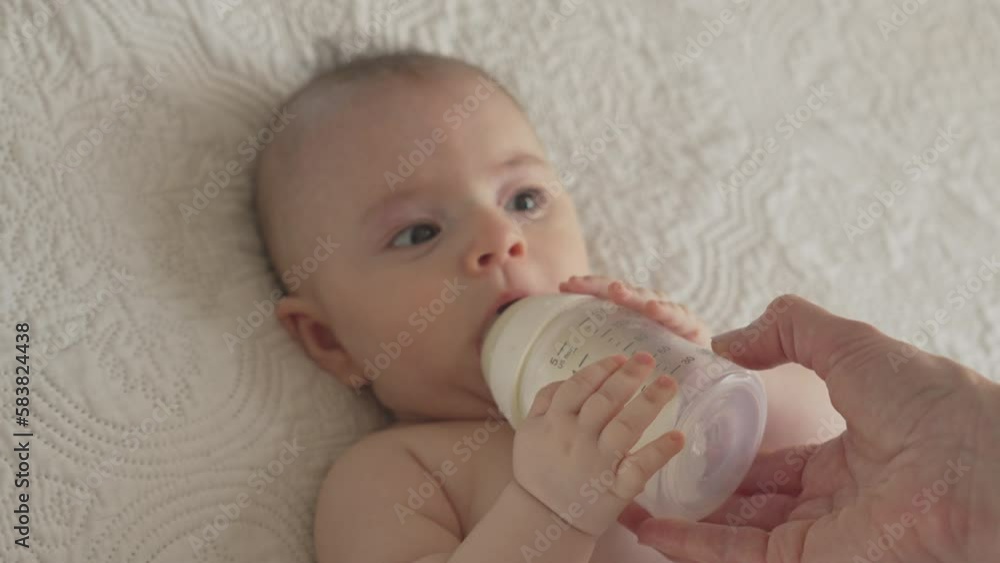 Baby sucking on milk bottle, newborn feeding time with mommy in white bright bedroom Baby sucking on milk bottle, newborn feeding time with mommy in white bright bedroom