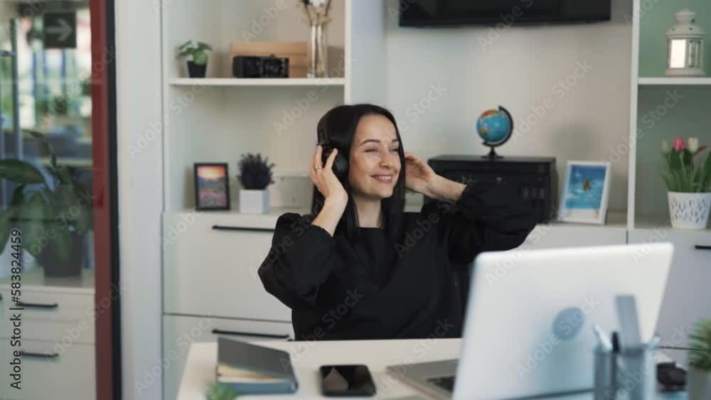 The beautiful middle-aged woman is sitting at a desk in her office in front of her laptop. She's listening to music on wireless headphones. The woman dances and waves her arms to the beat. The beautiful middle-aged woman is sitting at a desk in her office in front of her laptop. She's listening to music on wireless headphones. The woman dances and waves her arms to the beat.