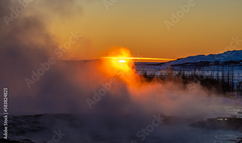 Iceland's great Strokkur geyser just before erupting with mist and smoke backlit with the orange sunset sun behind it