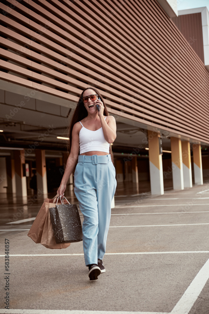 vertical photo of a caucasian woman holding shopping bags and calling with a mobile phone. Happy people enjoy sales day walking outdoors. Shopaholic Lady using cell and smiling