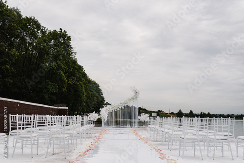 Setup wedding ceremony on pier. Wedding arch decorated with flowers and white chairs for guests. Wedding atmosphere, scenery, celebration. Photography, concept.